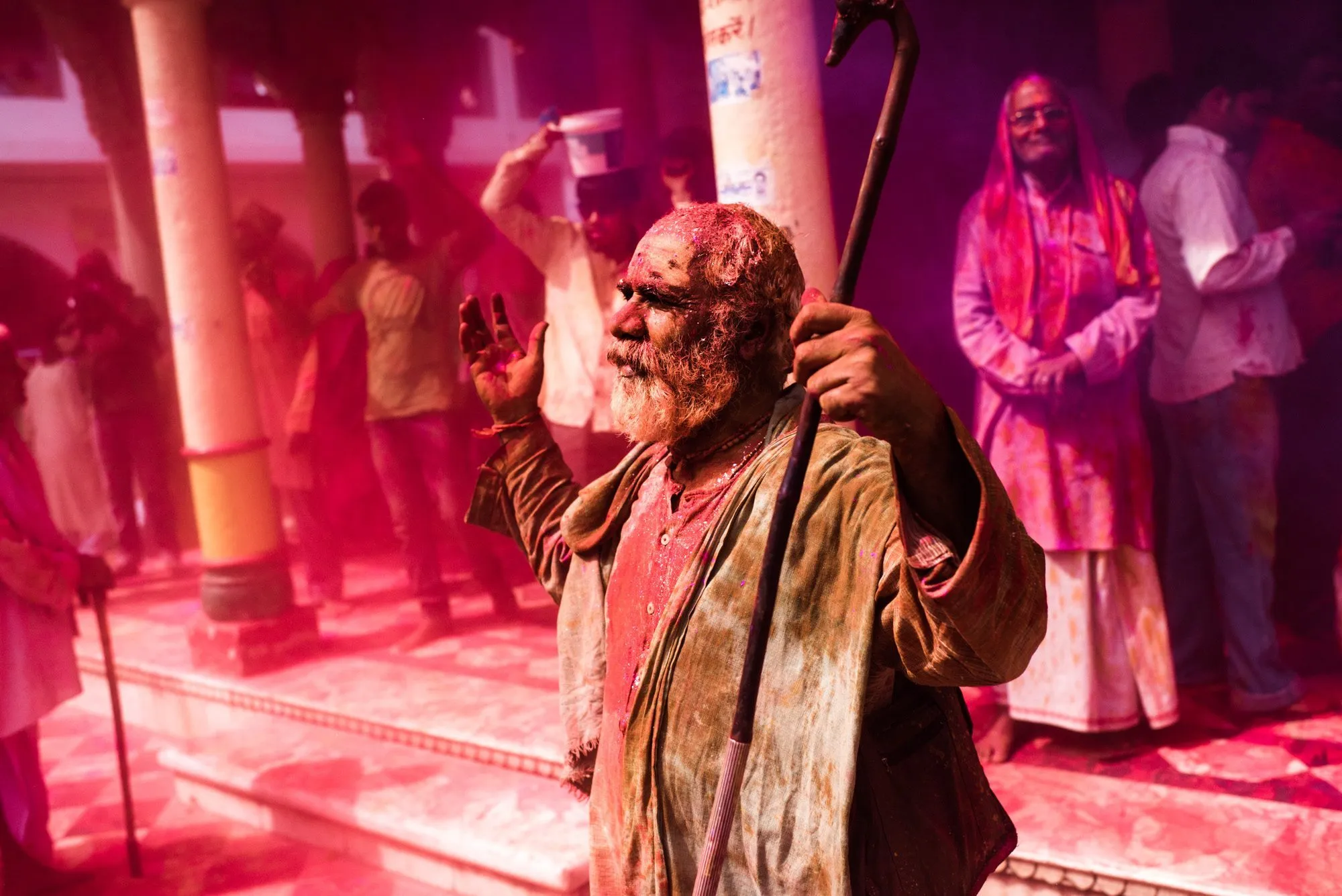 An elder coated in red and yellow powder holds a staff inside a temple courtyard during the Holi festival in India.