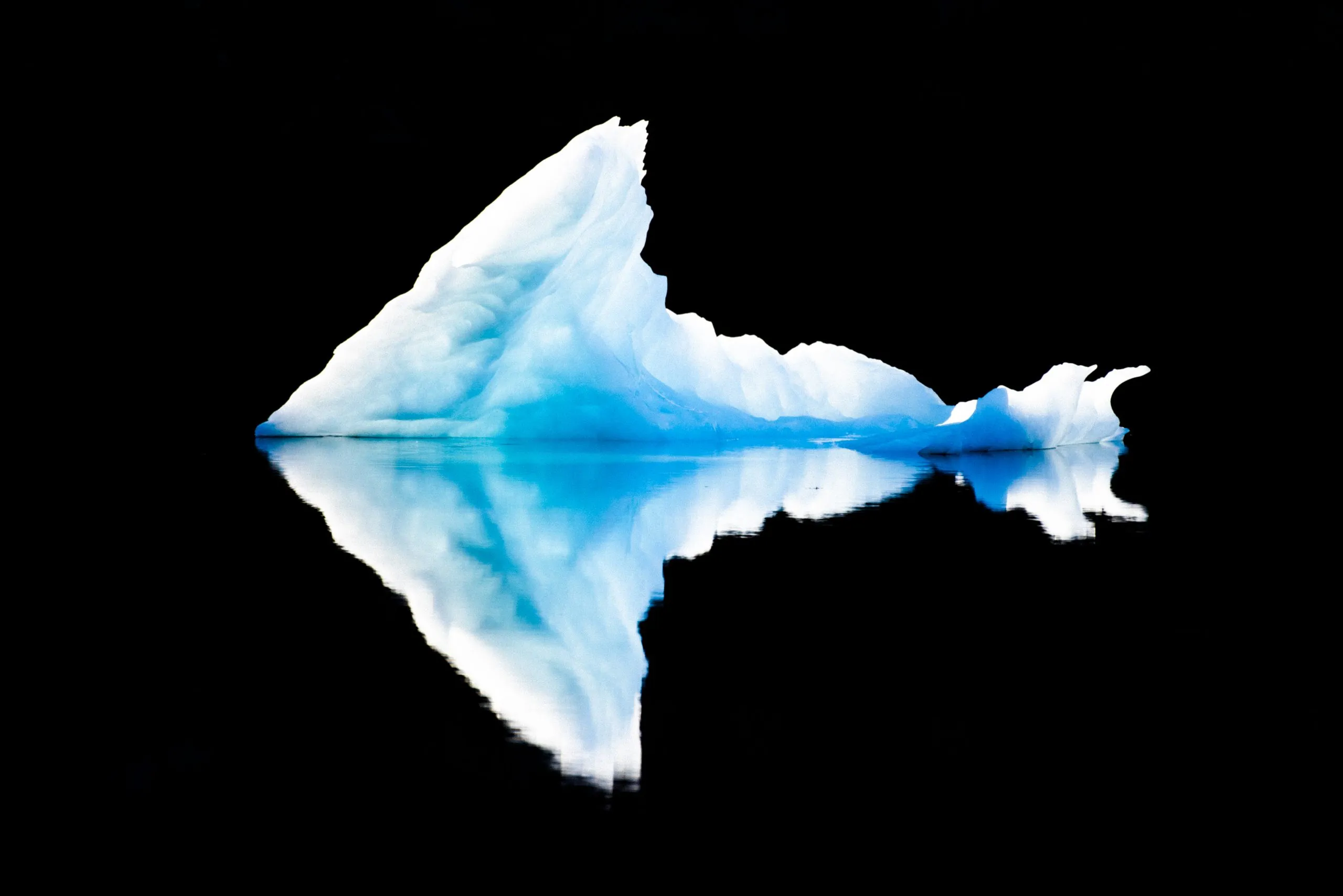 A glowing pyramid-shaped iceberg floats on a black lake-like sea, mirrored in the still water against an inky background.