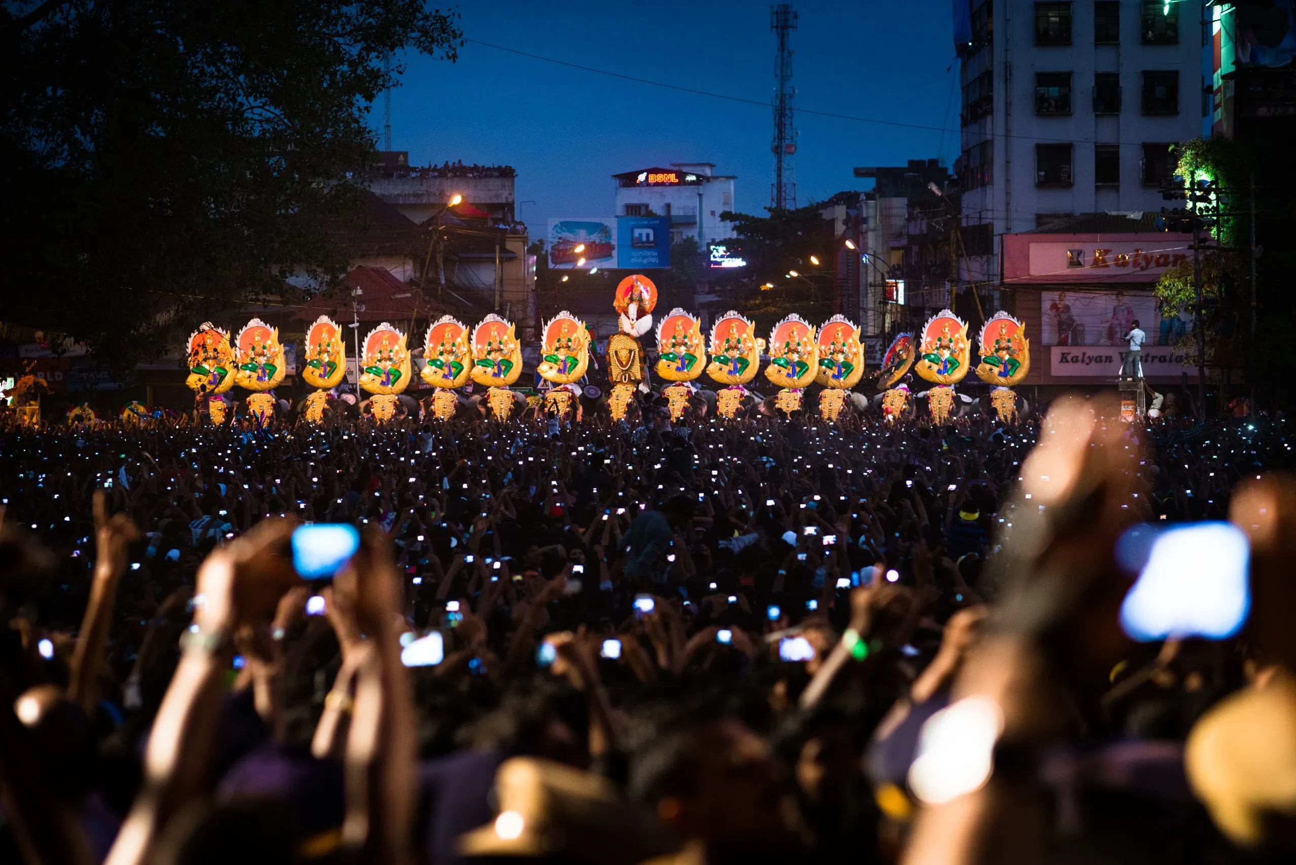 A vast nighttime crowd raises mobile phones to film a row of decorated elephants at the Thrissur Pooram festival in Kerala, India.