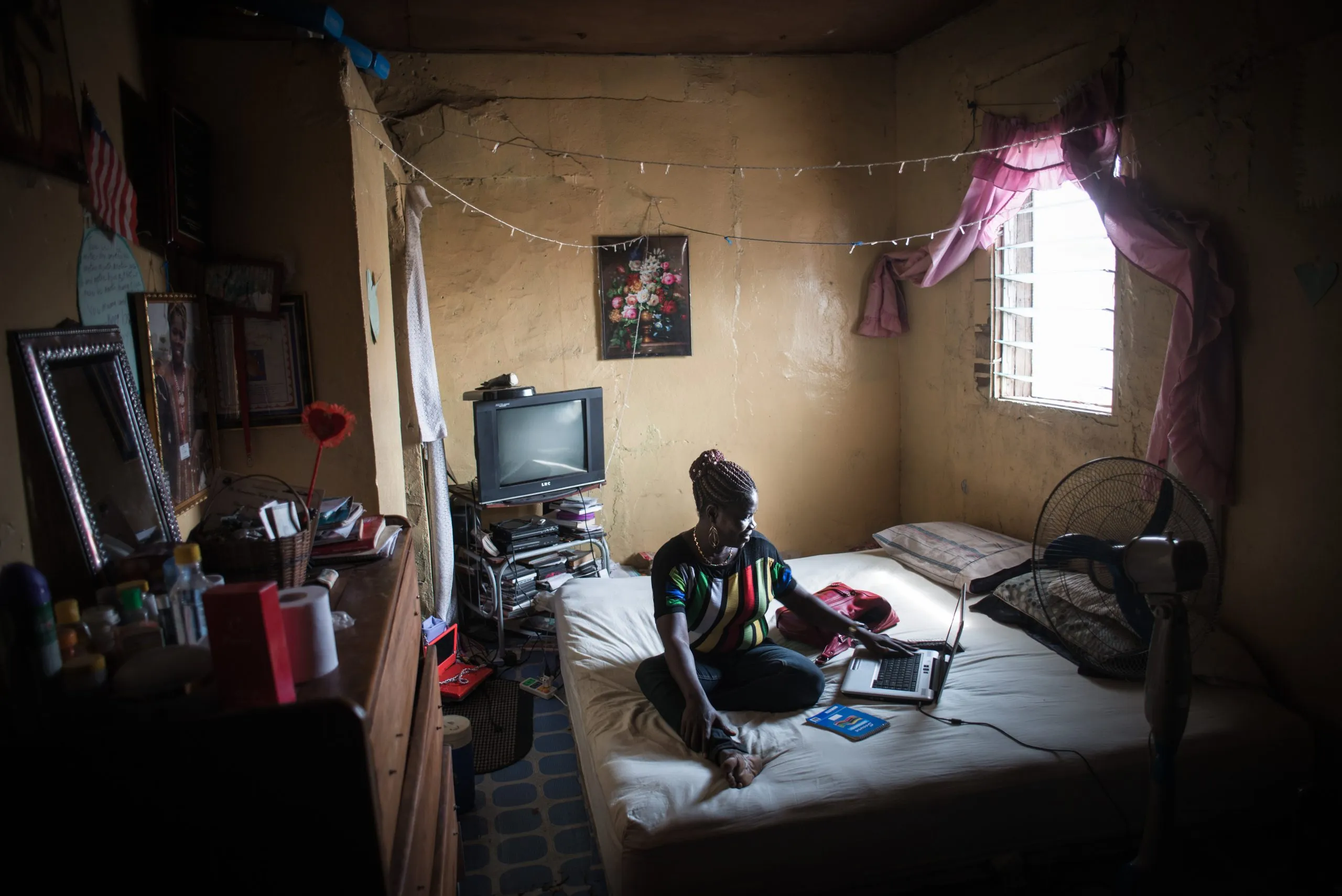 A Liberian woman sits on a bed in a small Monrovia home, working on a laptop, a US flag pinned to the wall behind her.