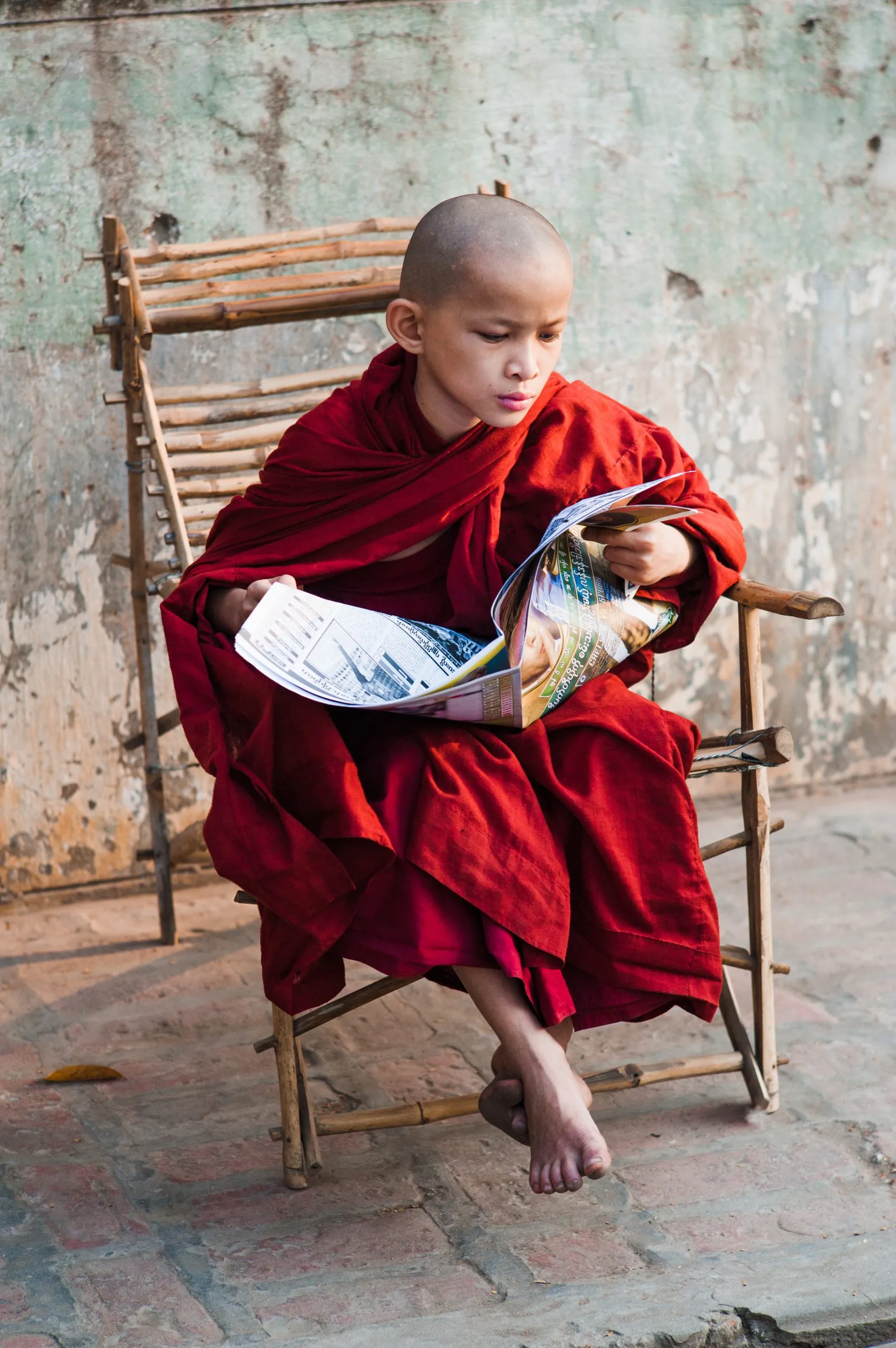 A young novice monk in red robes sits in a wicker chair on a Myanmar street, leaning forward to read a magazine.