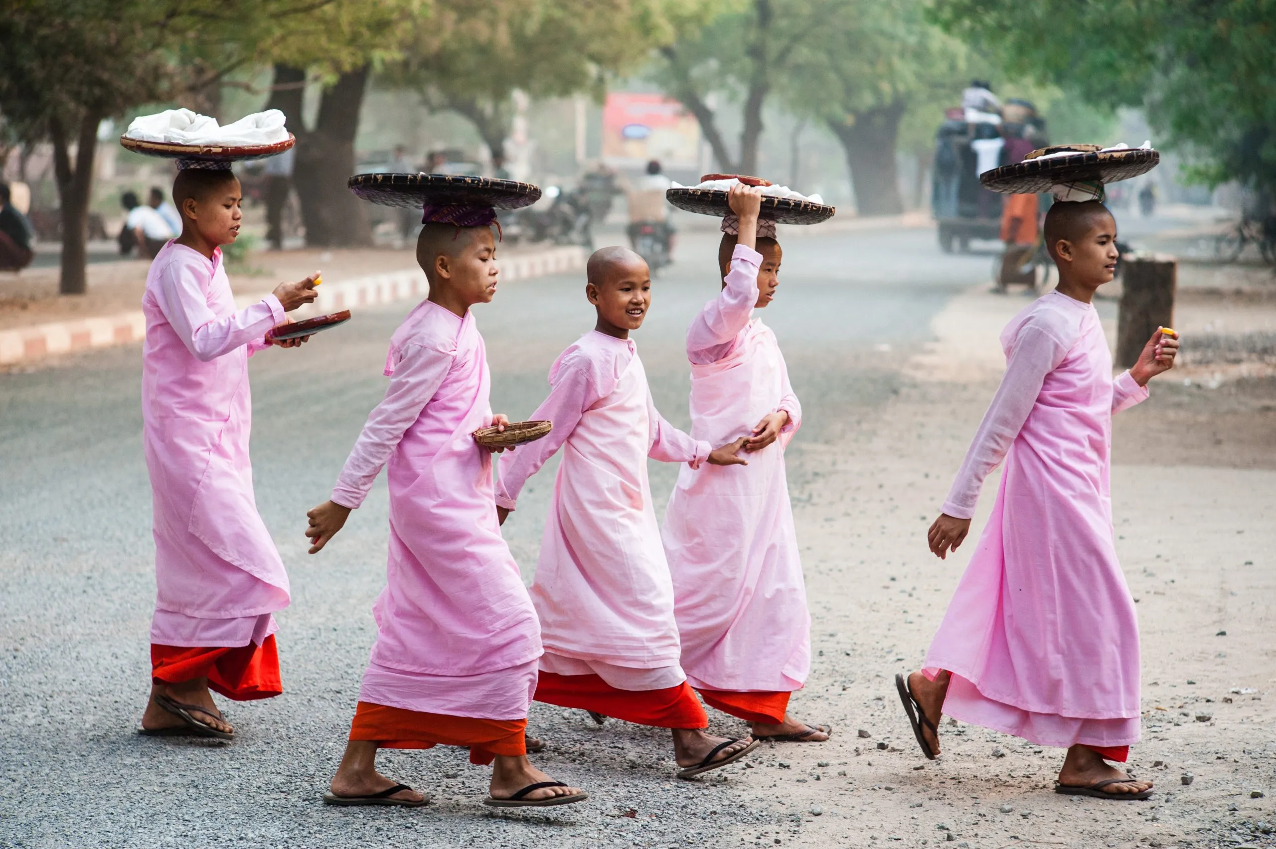 Five young Buddhist nuns in pink robes walk barefoot along a road in Bagan, Myanmar, balancing alms baskets on their heads.