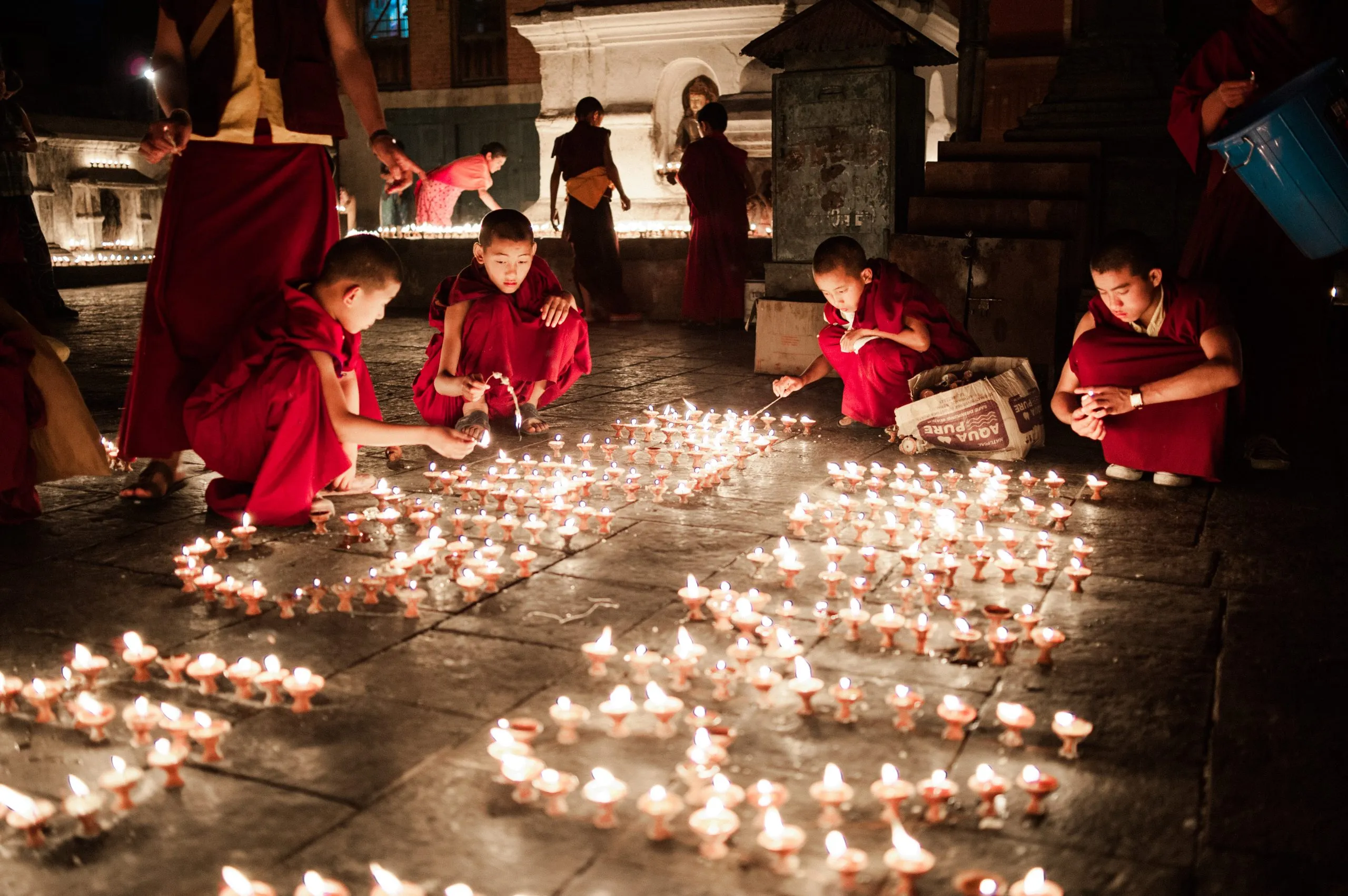 Several young monks light hundreds of butter lamps arranged in a glowing pattern on the courtyard of Boudhanath stupa in Kathmandu.