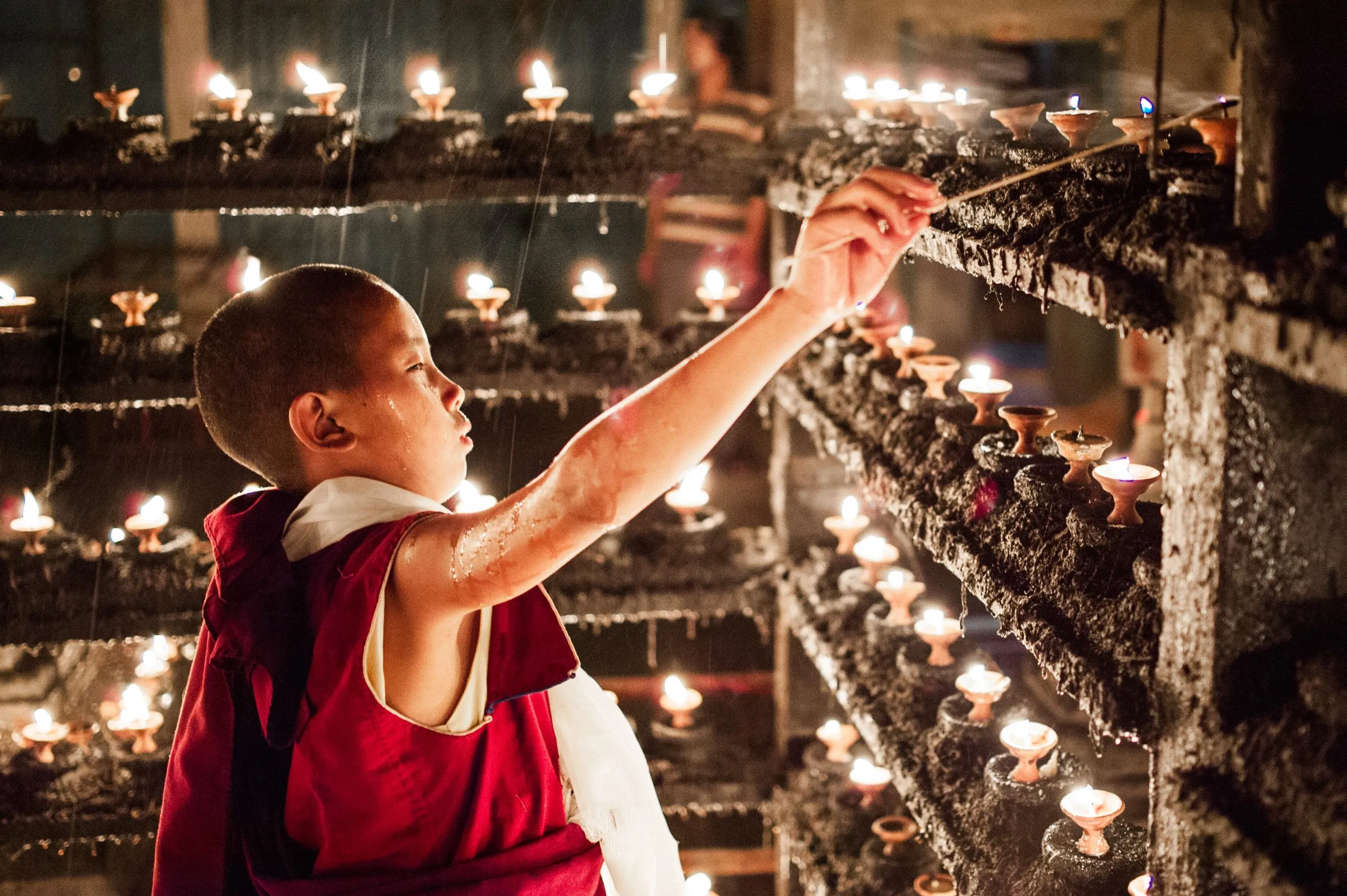 A young Nepali Buddhist novice lights a row of butter lamps along a candle-encrusted wooden rack in a Kathmandu monastery.